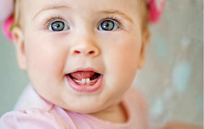 A close up of a baby girl wearing pink with big blue eyes, holding her mouth open showing her first two teeth