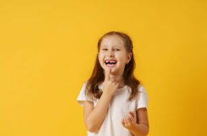 A young girl smiling and point to her missing teeth while smiling against a yellow background.