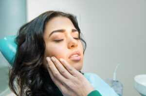 A woman sitting on a dental chair at the dentist's office and she is holding the right side of her jaw due to pain from her mouth