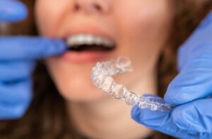 An orthodontist wearing blue latex gloves inserts a finger into a patient's mouth while holding an Invisalign retainer.