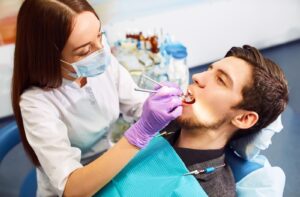 Female dentist or dental hygienist using metal tools to clean male patient's teeth.